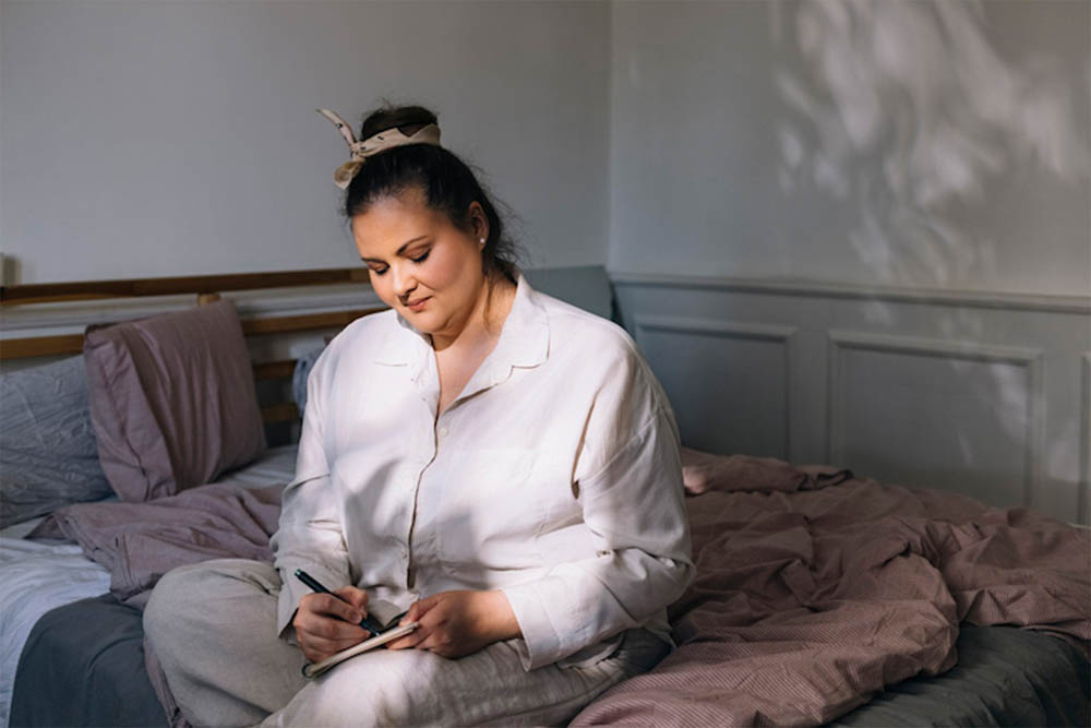 A woman sitting on her bed in soft morning light, writing in a journal while wearing comfortable sleepwear.