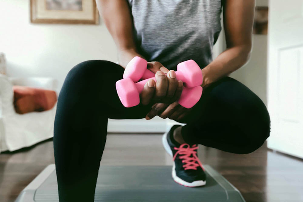 Person doing a squat exercise at home while holding two pink dumbbells, wearing athletic clothes and sneakers.