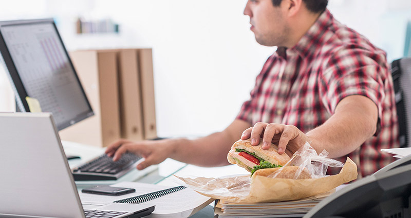 A man in a checkered shirt sitting at a desk with a computer, reaching for a sandwich wrapped in paper