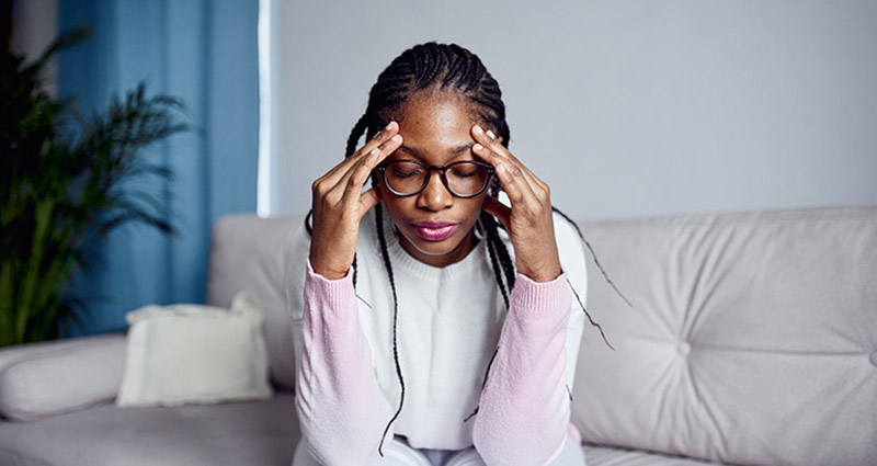 A woman sitting on a couch with her hands on her temples, looking stressed or overwhelmed