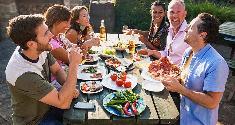 A group of six people enjoying a lively outdoor meal at a picnic table, filled with a variety of dishes. 