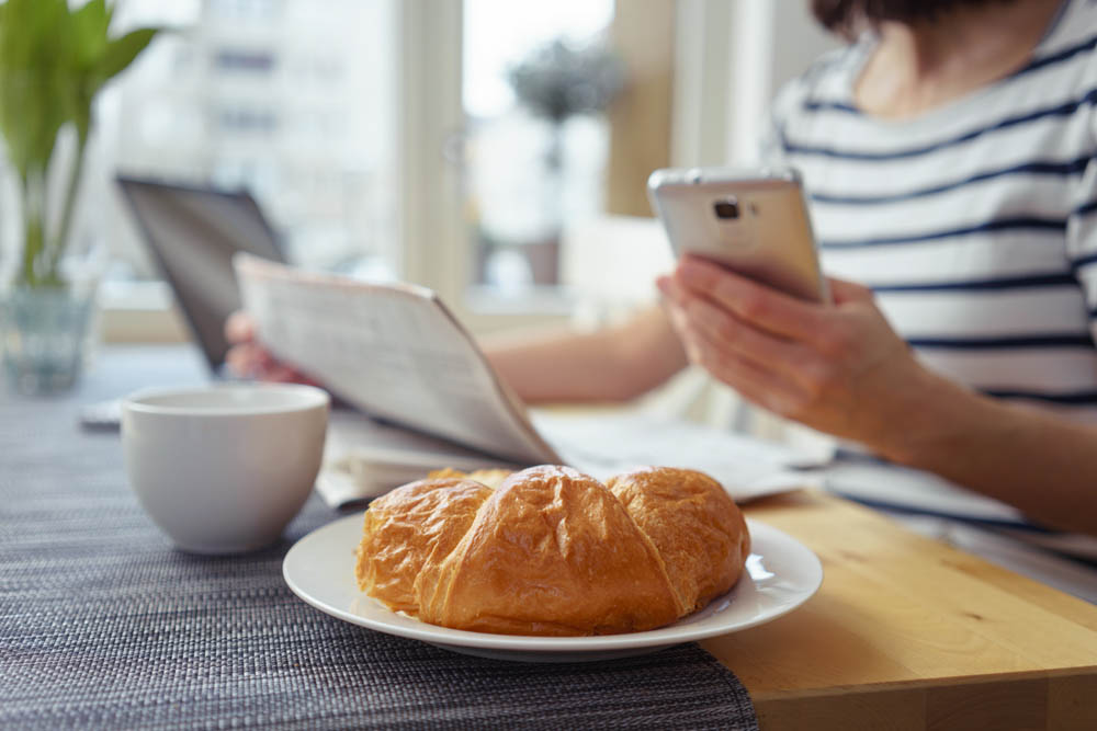 The woman is sitting with the daily newspaper and her phone at breakfast