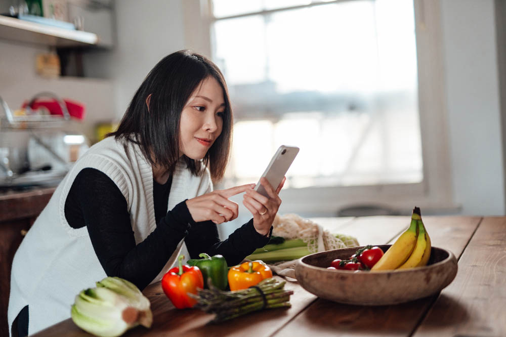 Woman in a kitchen using a smartphone near a wooden table with assorted vegetables and bananas.