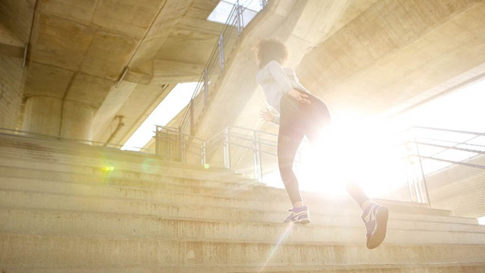 Person running up outdoor stairs with bright sunlight in the background, wearing athletic clothing and shoes.