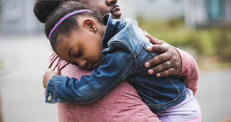 A tender moment between an adult and a young girl, with the girl resting her head on the adult's shoulder, appearing calm and content.