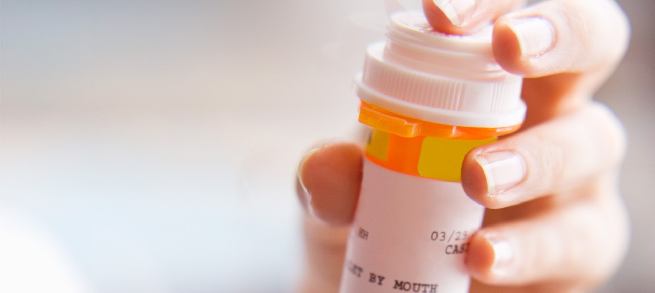 A close-up of a person's hand holding a prescription medication bottle, with the cap partially unscrewed