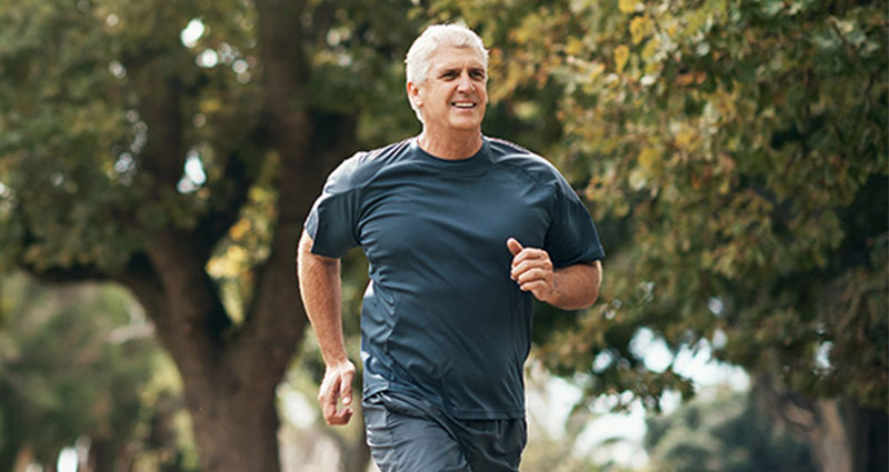 A man jogging outdoors in a park, wearing a dark blue athletic shirt and shorts