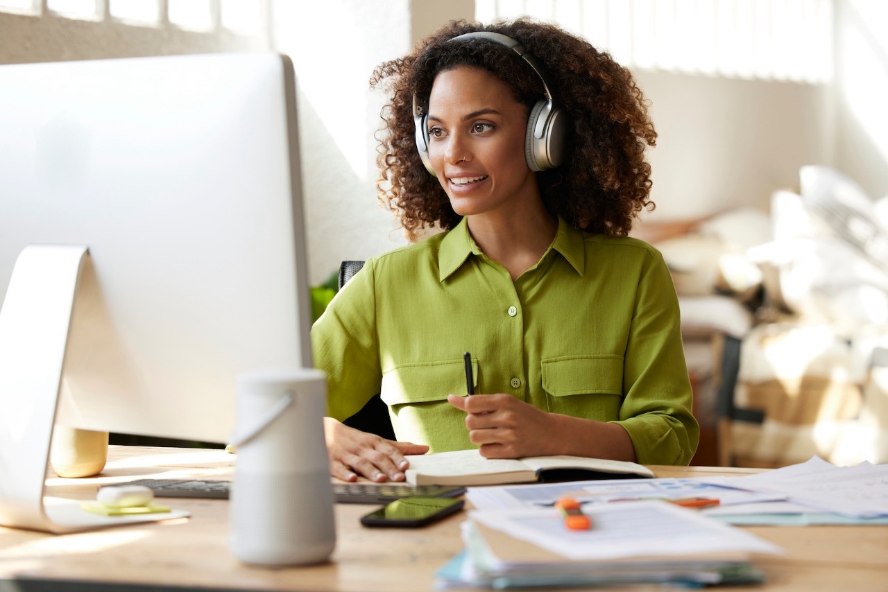 Smiling woman wearing headphones, sitting at a desk with a computer, taking notes while attending an online coaching session.