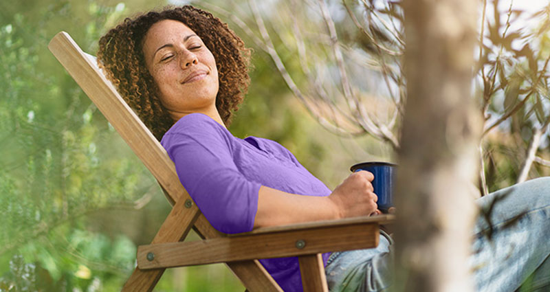 A woman sitting comfortably in a wooden chair outdoors, holding a mug and enjoying the serene environment