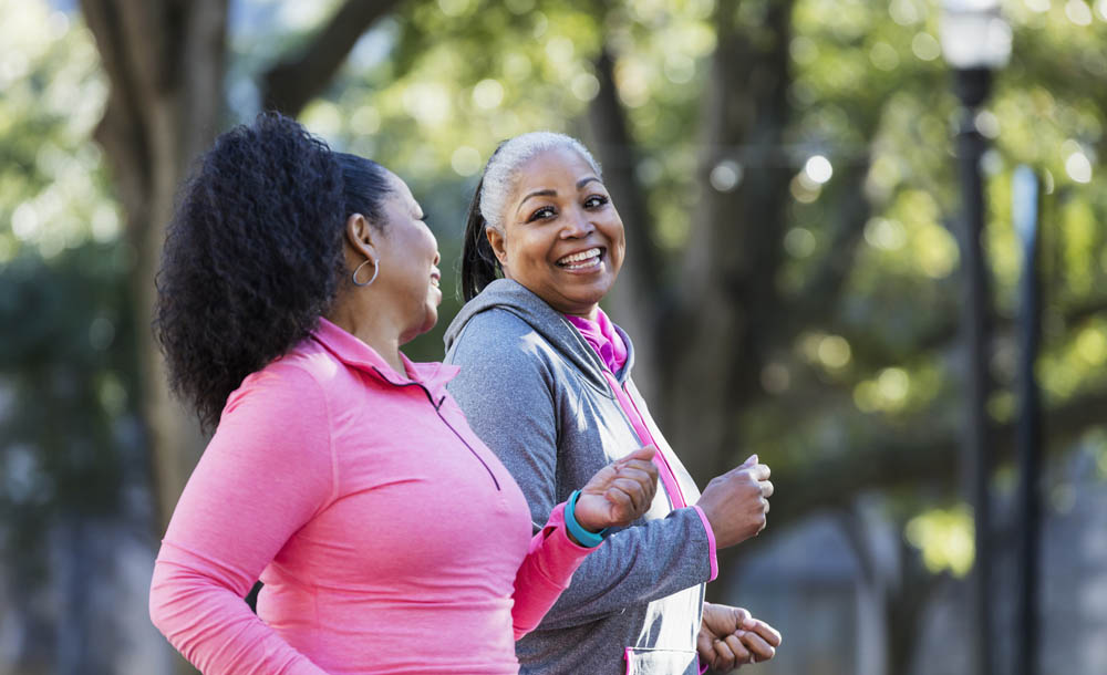 Two African-American women exercising together in the city, jogging or power walking, laughing and conversing. Buildings and trees are out of focus in the background. The one in pink is in her 60s and her friend is in her 50s.