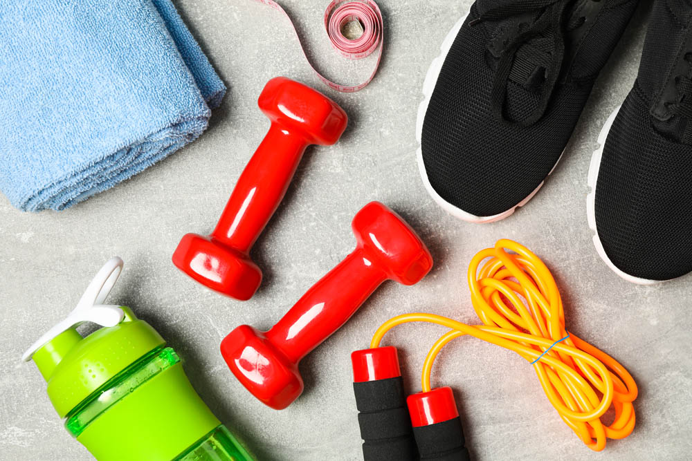 Fitness items including red dumbbells, a blue towel, black sneakers, a green water bottle, a measuring tape, and an orange jump rope on a gray surface.