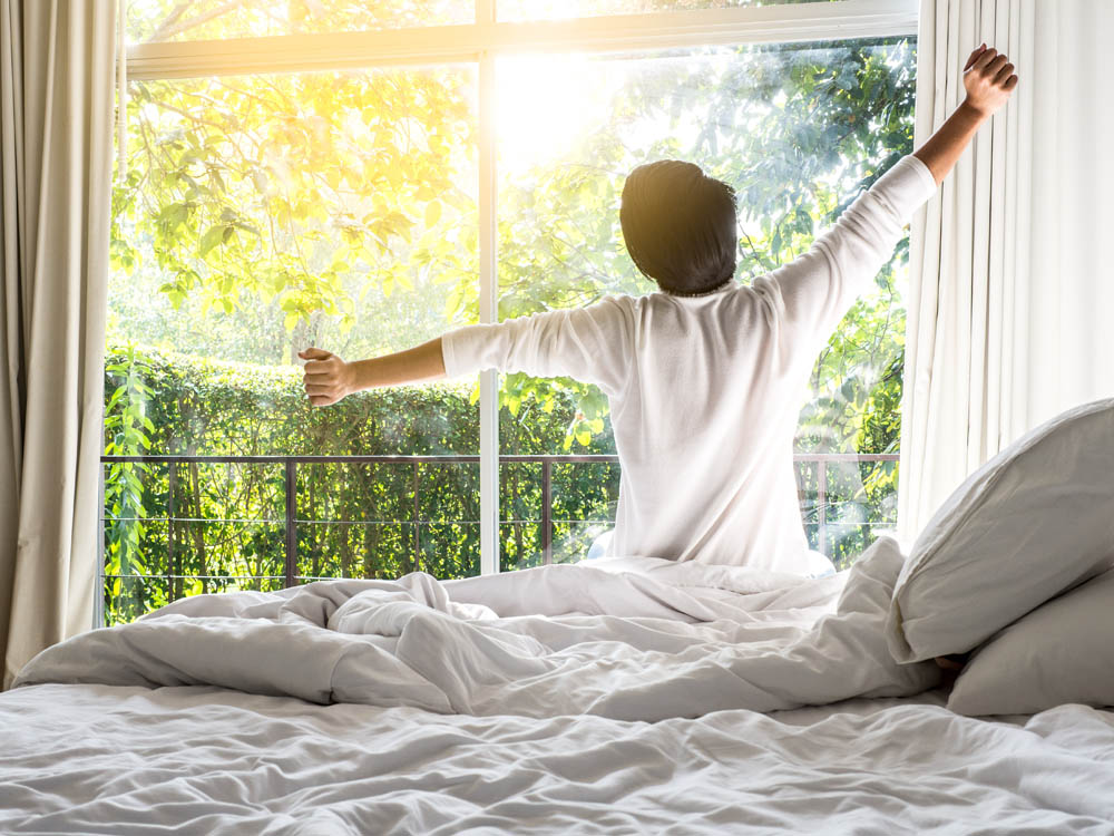Person stretching arms while sitting on a bed, facing a sunlit window with green foliage outside.