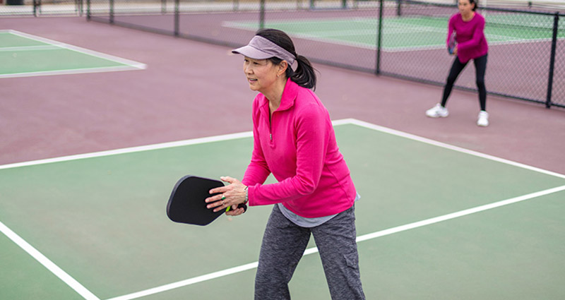 A woman in a pink long-sleeve shirt and visor preparing to play pickleball on a court