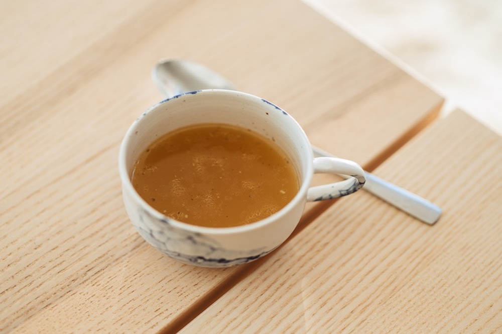 A small cup filled with brown liquid sits on a wooden table, accompanied by a metal spoon resting beside it. The cup has a marble design.