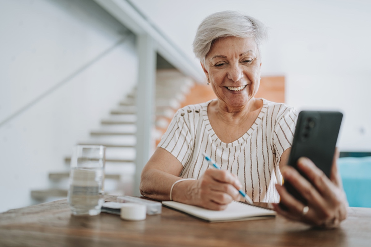 Smiling older woman writing in a notebook while looking at her phone.