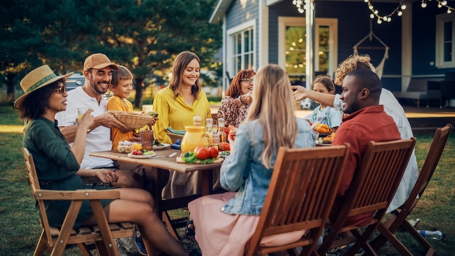 A group of people sitting around an outdoor dining table, enjoying a meal. The table is set with food and drinks, and there are hanging lights nearby.