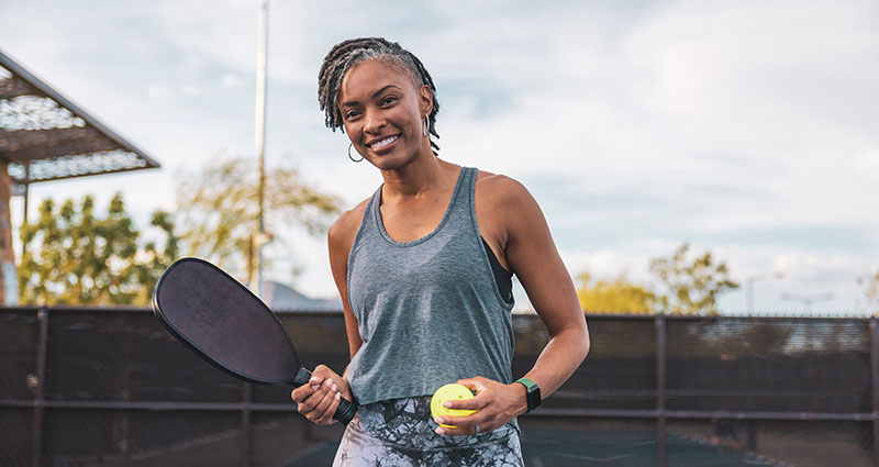 A smiling woman holding a tennis racket and a tennis ball, standing on a tennis court. She is wearing a sleeveless top and has her hair styled in a braid, with a cloudy sky in the background.