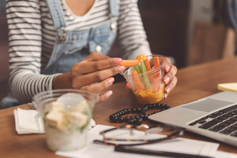 A person in striped sleeves and denim overalls dipping a carrot stick into hummus while working on a laptop.