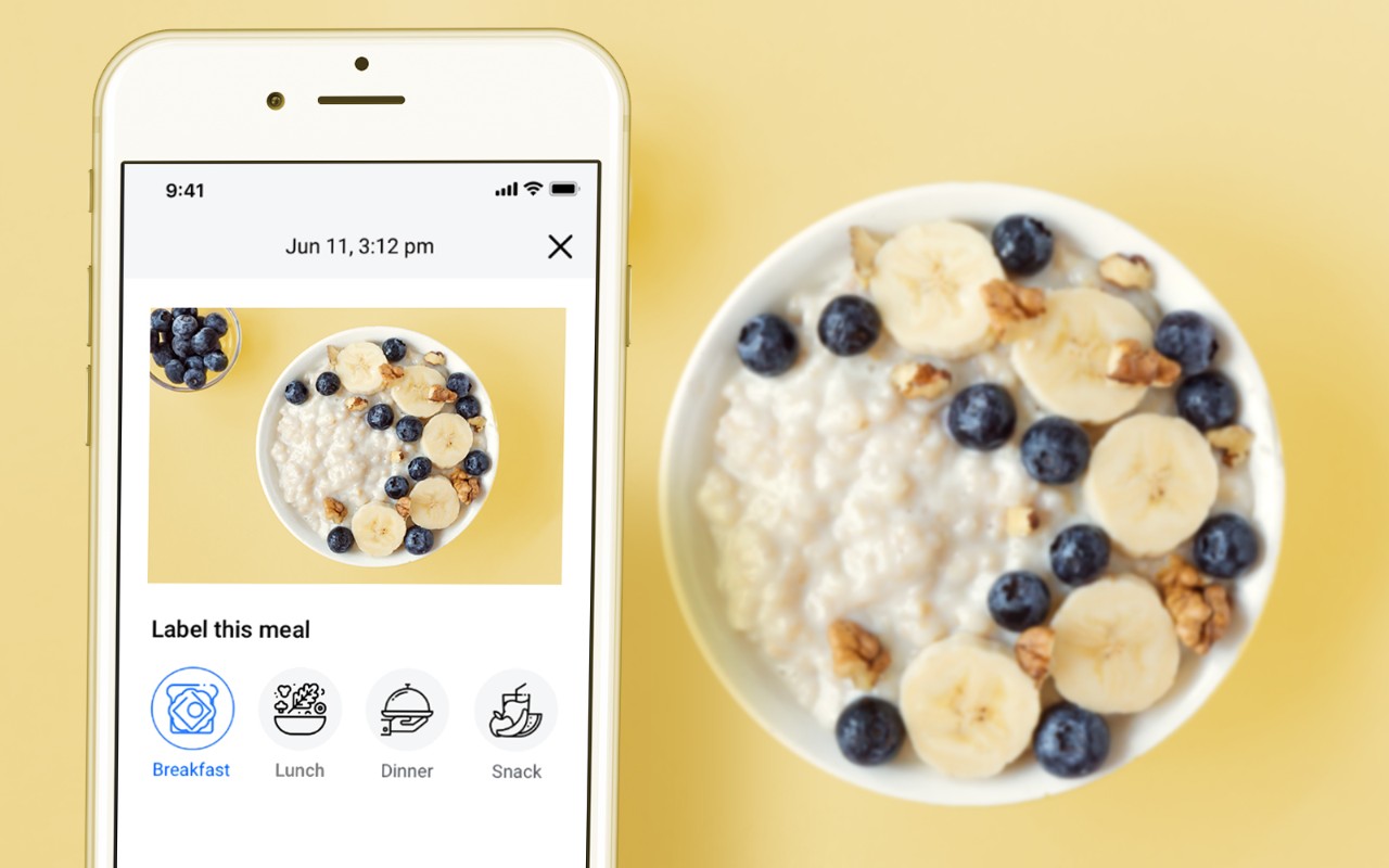 A bowl of cottage cheese topped with banana slices, blueberries, and nuts next to a smartphone displaying a photo of the same meal.
