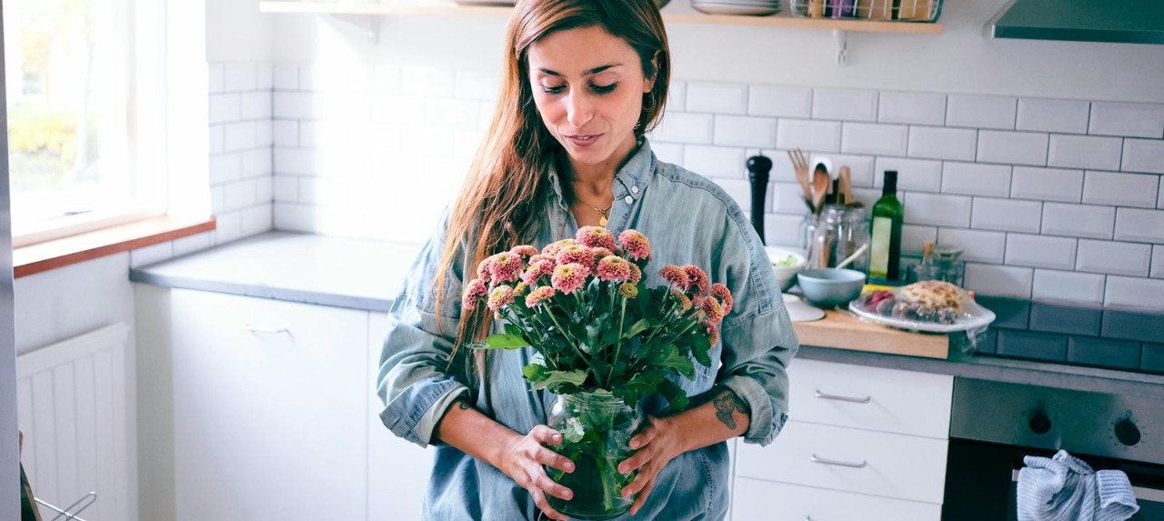 A woman in a cozy kitchen arranging a bouquet of flowers in a glass jar.