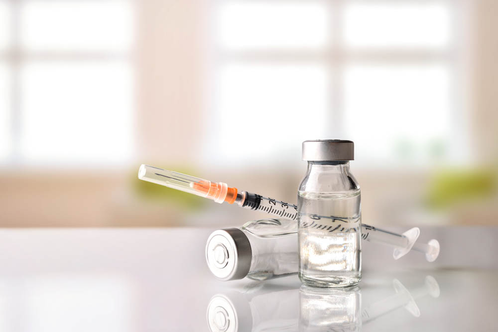 Two vaccine vials and a syringe with a needle placed on a reflective white surface.