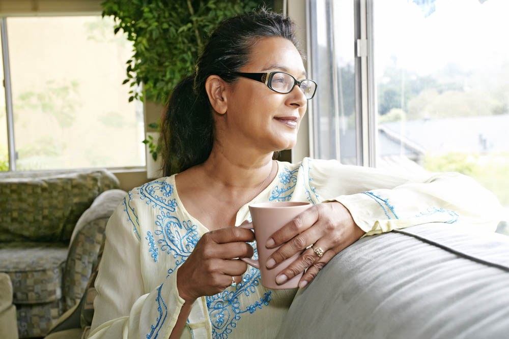 Woman with glasses holding a mug, looking out a window while sitting on a sofa in a light-filled room.