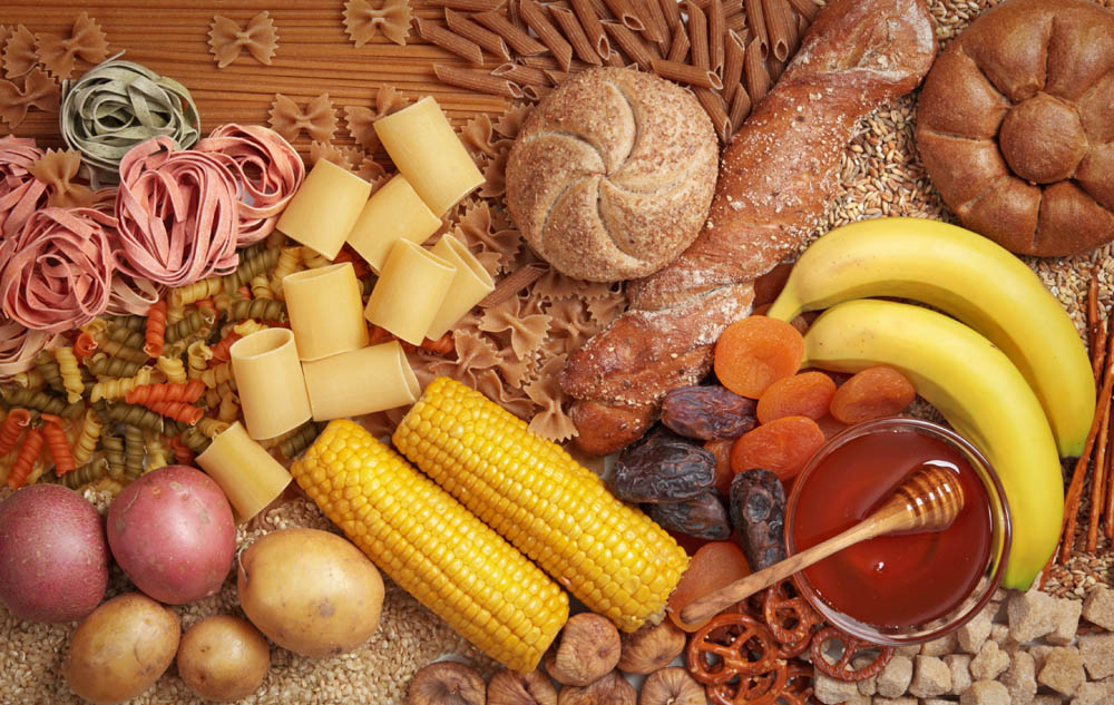 Assorted foods including pasta, bread, bananas, corn, dried fruits, honey, and potatoes arranged together on a wooden surface.