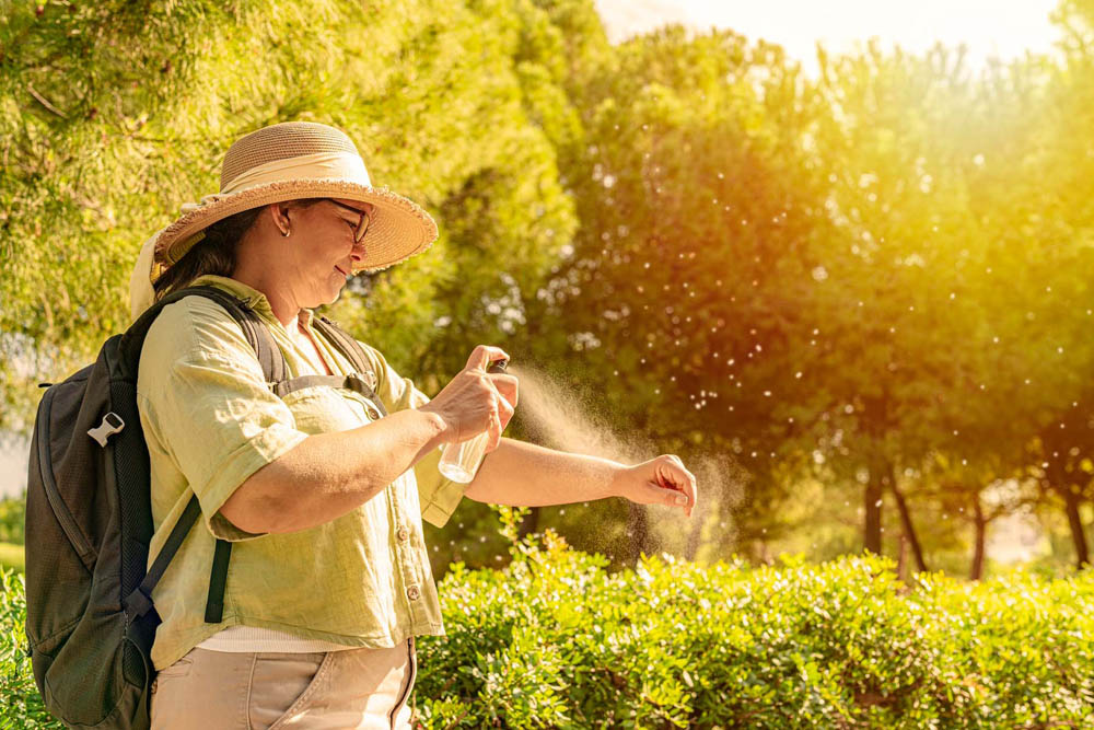 A person in a hat and sunglasses sprays insect repellent on their arm while standing in a sunny park, with trees in the background.