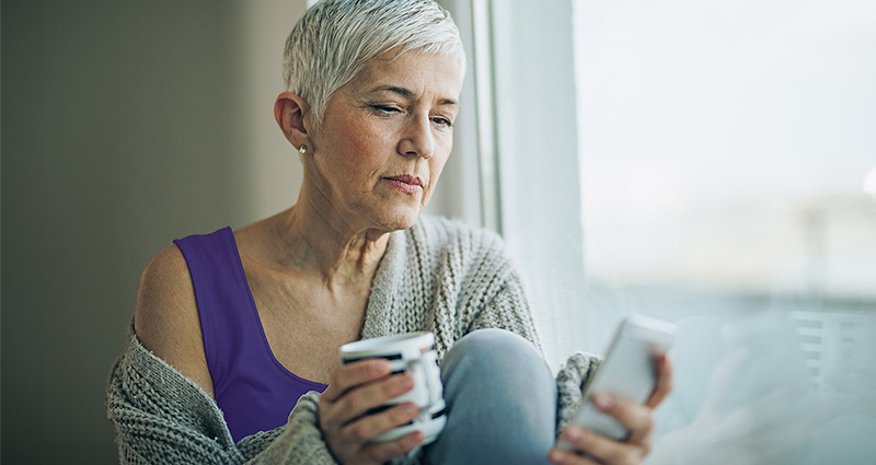 A mature woman sitting by a window, holding a cup and looking thoughtfully at her phone.