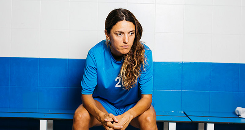 A female soccer player in a blue jersey sitting on a bench, looking serious and focused