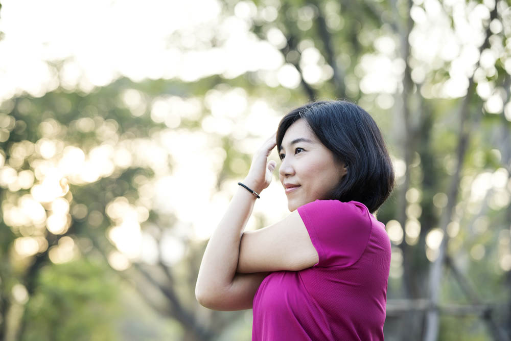 Warm Up and Cool Down Stretching Exercises. Cheerful Asian female runner doing shoulder stretche before morning jogging on trails in public park.