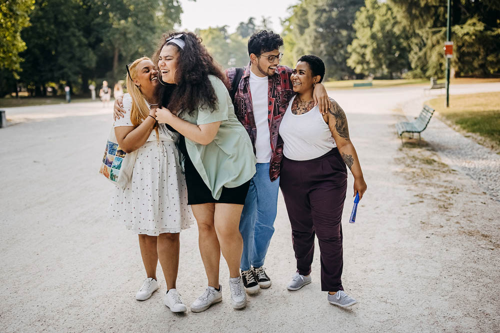 Four friends walking on a park path, laughing and embracing. Trees and benches line the background.