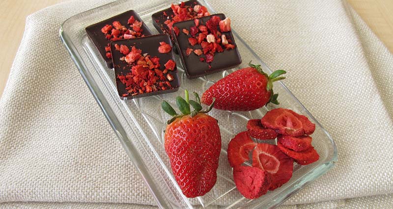 A glass plate holding dark chocolate squares topped with freeze-dried strawberry pieces, accompanied by fresh strawberries and sliced dried strawberries, resting on a textured beige surface.