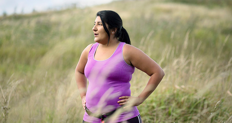 A woman in a purple tank top standing outdoors, with her hands on her hips and looking off to the side