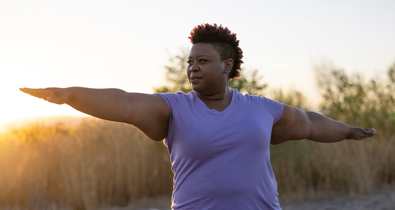 A woman in a purple shirt performing a yoga pose outdoors, with her arms extended