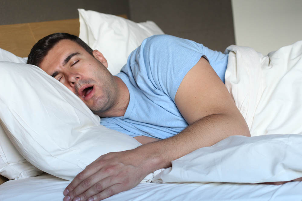 Man in a blue T-shirt sleeping on his side with his mouth open, head on a pillow, under white bedding.
