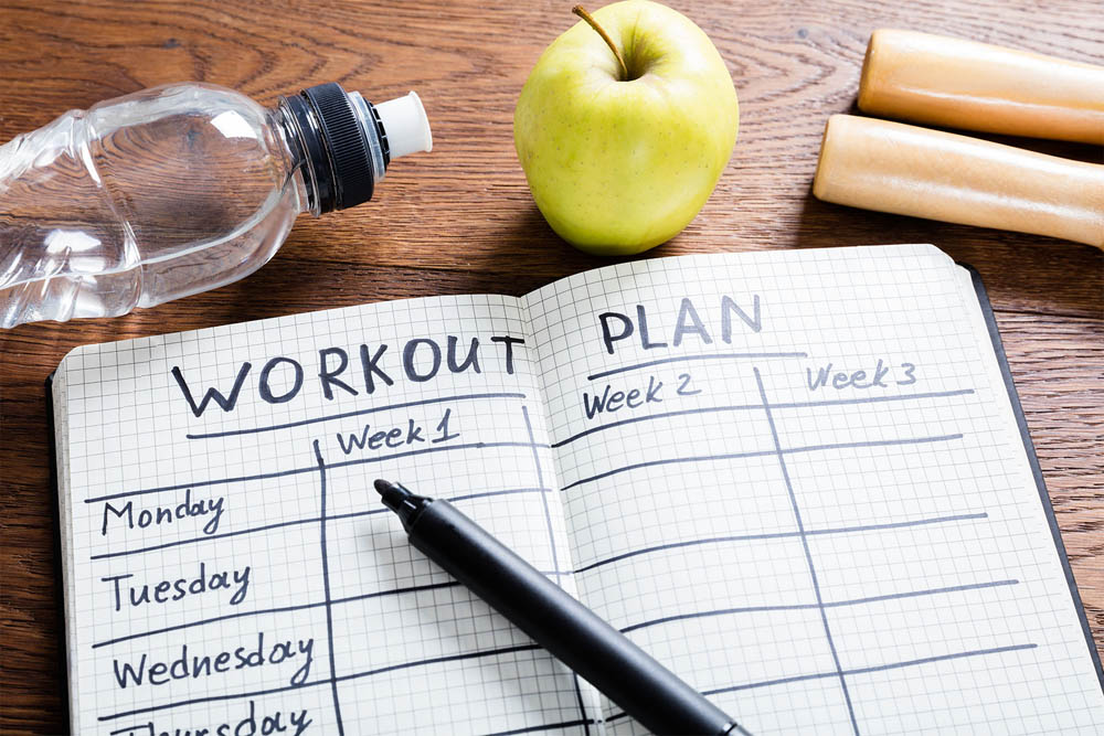 Open workout planner on a table with a pen, water bottle, green apple, and two wooden jump rope handles.