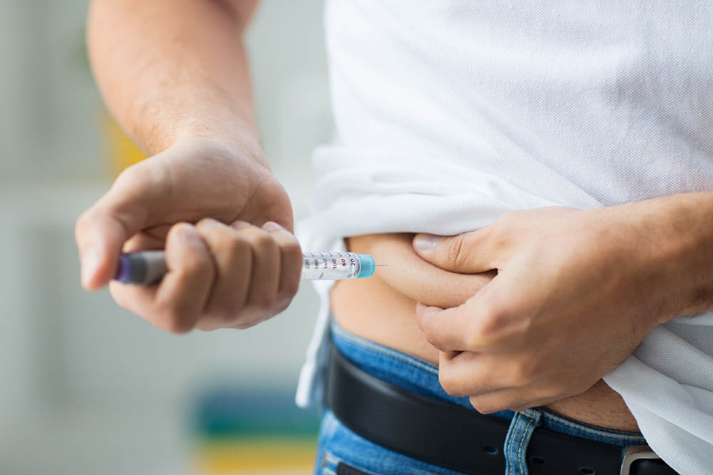 Person injecting insulin into their abdomen using a syringe pen, lifting their white shirt slightly, standing in a well-lit room.
