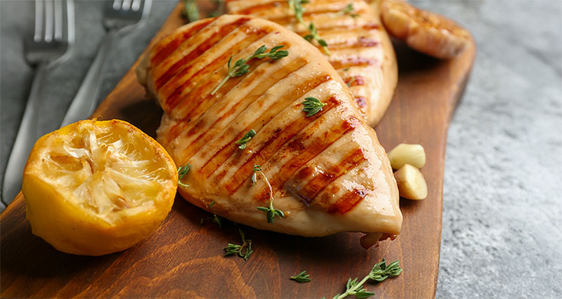 Grilled chicken breasts on a wooden board with fresh herbs, lemon halves, and garlic cloves. Two forks are in the background.