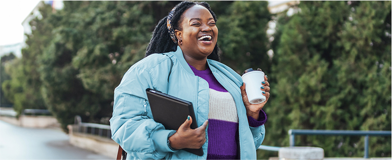 A happy, smiling woman walks outside, carrying a tablet and a thermos.