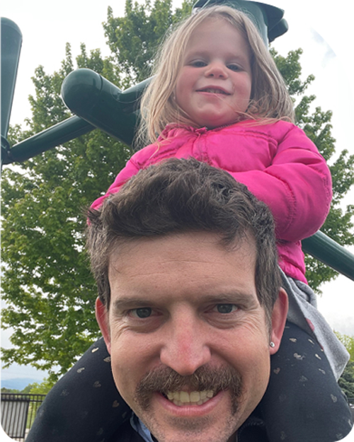  Smiling man with a mustache carrying a young girl on his shoulders at a playground