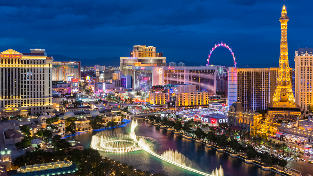 A vibrant nighttime aerial view of the Las Vegas Strip, showcasing illuminated buildings, including a replica of the Eiffel Tower and the Ferris wheel, with colorful lights reflecting on the water of a nearby fountain.
