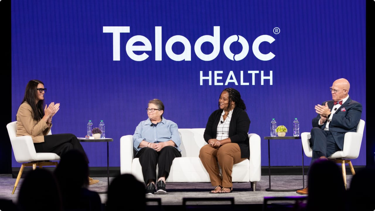 Four professionally dressed panelists sit on stage in white chairs having a discussion during Forum 2025. A panel behind them displays a Teladoc Health logo on a purple background.