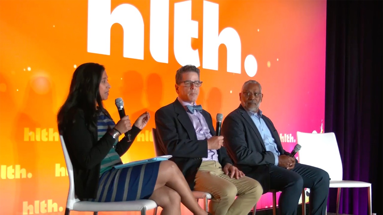 Three professionally dressed panelists sit on stage in white chairs having a discussion during HLTH. A large screen behind them displays the event name in white letters with orange background.