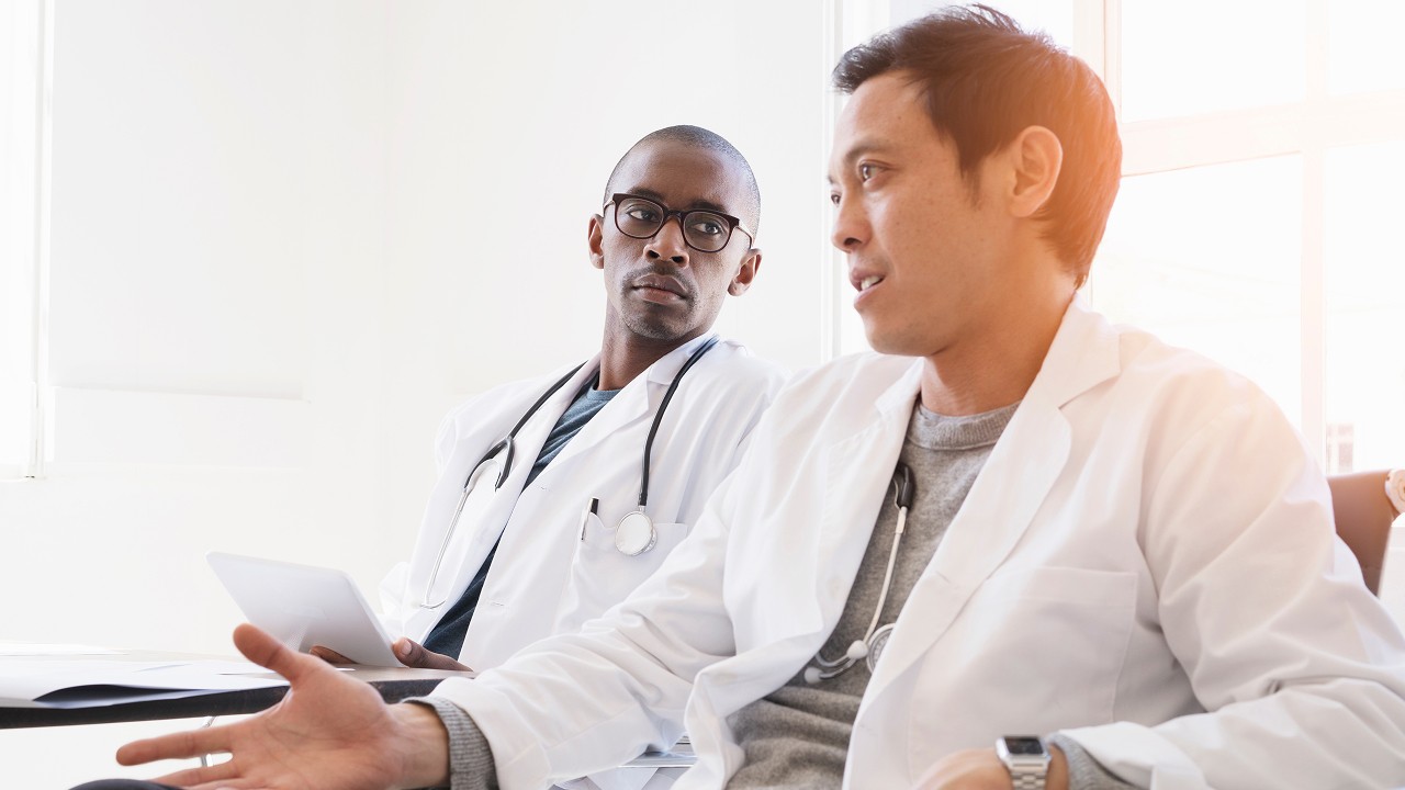 Two male doctors in white lab coats having a serious discussion in a bright room. One holds a tablet while the other gestures with his hand.