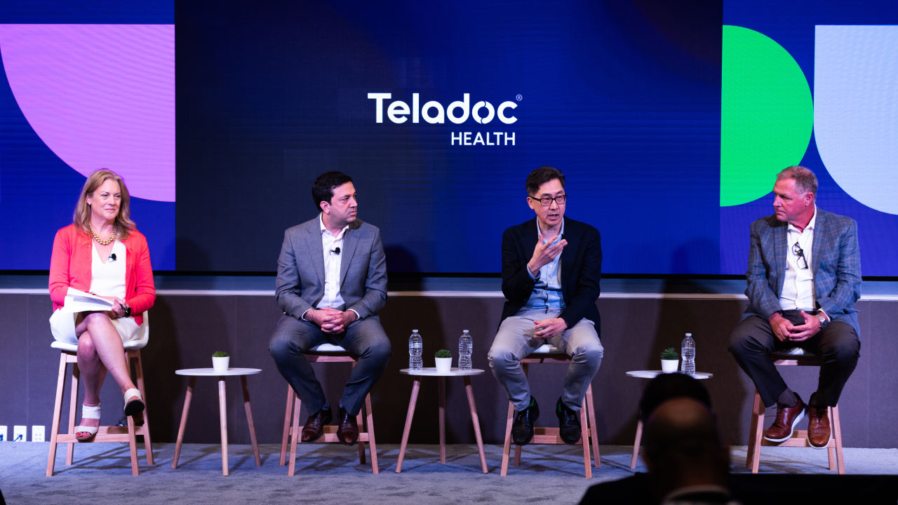 Four speakers seated on a stage during a Teladoc Health event, participating in a panel discussion