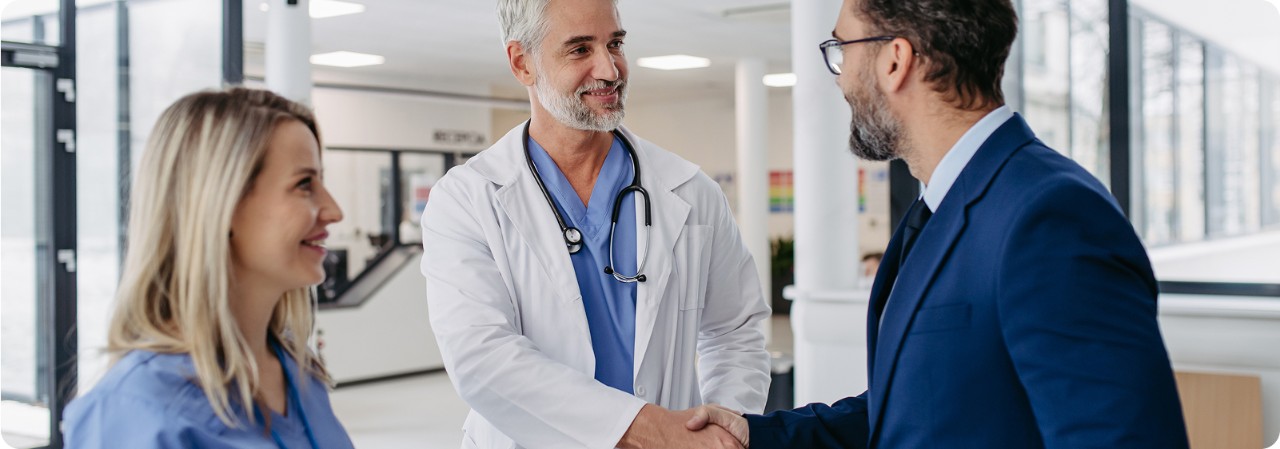Smiling male doctor in a white coat and stethoscope shaking hands with a man in a business suit, while a female health professional looks on in a modern medical facility.