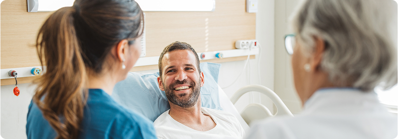 Smiling male patient sitting in a hospital bed, talking with two healthcare professionals during a hospital visit.