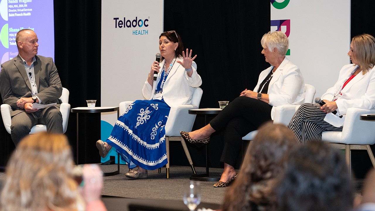 Four professionally dressed panelists sit on stage in white chairs having a discussion during Forum 2025. A panel behind them displays a Teladoc Health logo on a white background.