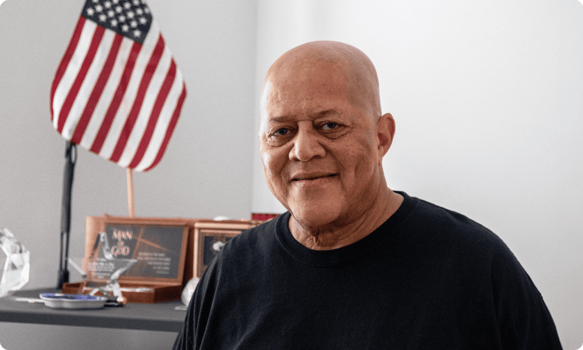 Person in a black shirt standing indoors with an American flag and plaques on a shelf in the background.